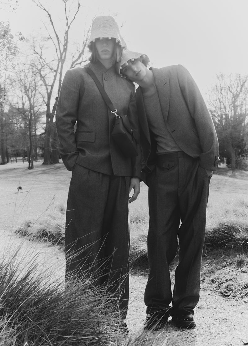Two men pose outdoors wearing suits and bucket hats. One man has his head resting on the shoulder of the other. Trees are visible in the background.