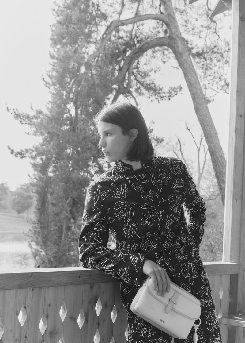 Monochrome portrait of a young woman in a patterned coat, leaning against a porch railing with trees visible in the background.