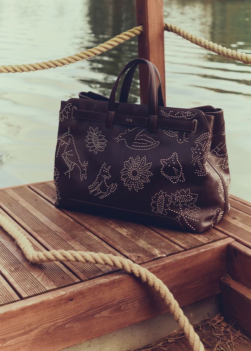 Brown leather tote bag with white floral and animal designs displayed on a wooden dock near a body of water with a rope border.
