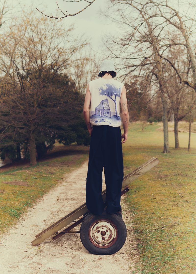 Person wearing a sleeveless shirt with a house design stands on a tire attached to a wooden plank in a grassy outdoor setting.