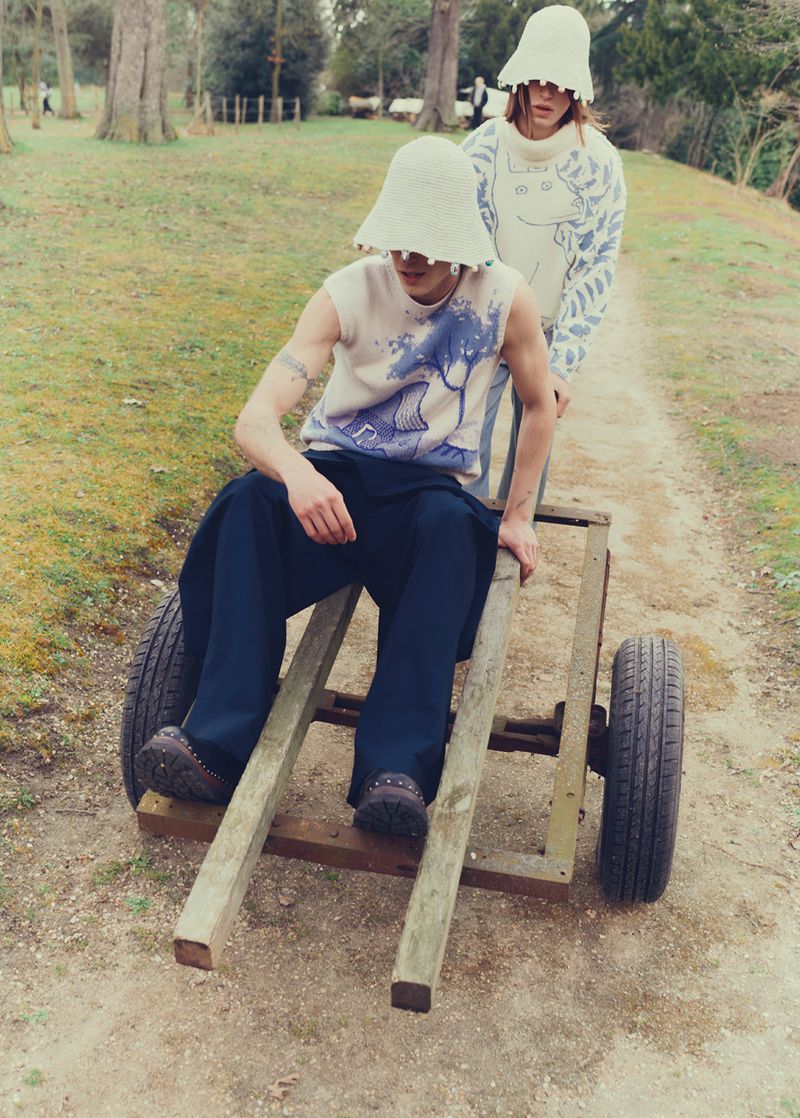 Two people in an outdoor setting with one man sitting on a wooden cart being pushed by another, both wearing bucket hats.