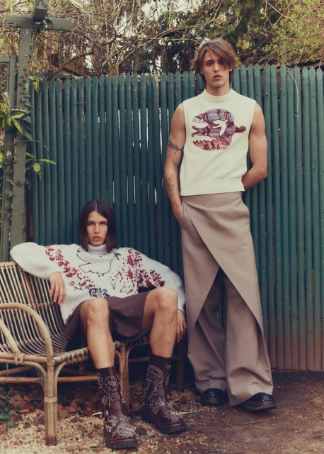 Two young adult men pose outdoors against a green metal fence; one is seated on a rattan chair, wearing a sweater and boots.