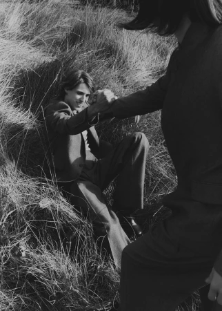 Black-and-white outdoor shot of a man in a suit sitting in tall grass as another person reaches out to help him up.