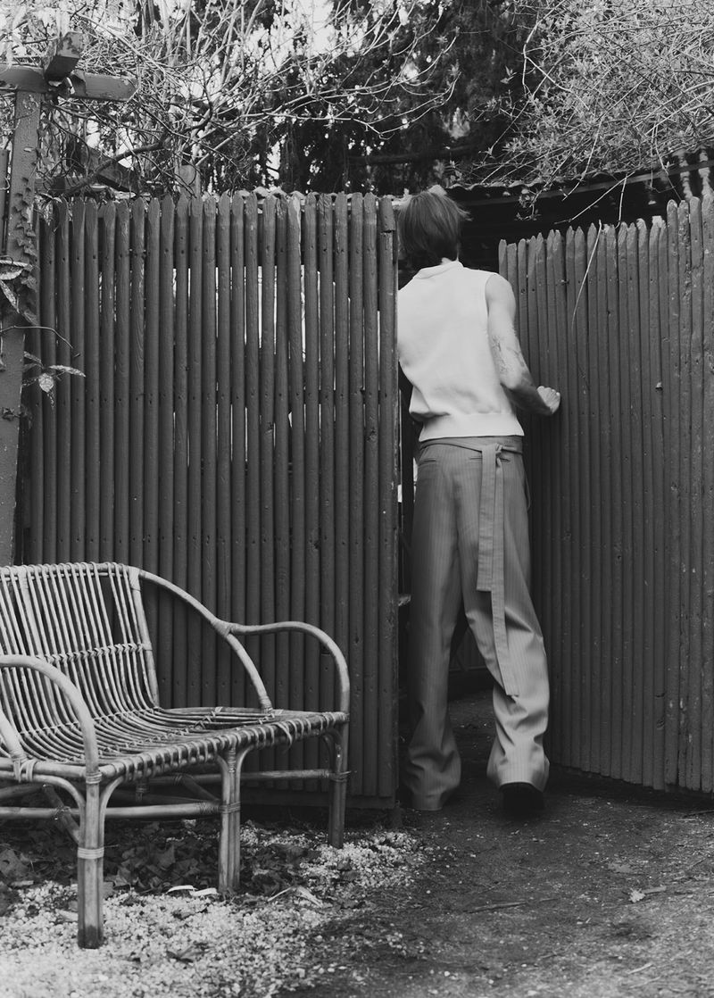 Monochrome image of a person opening a weathered wooden gate in a garden, featuring a wicker chair and lush foliage.