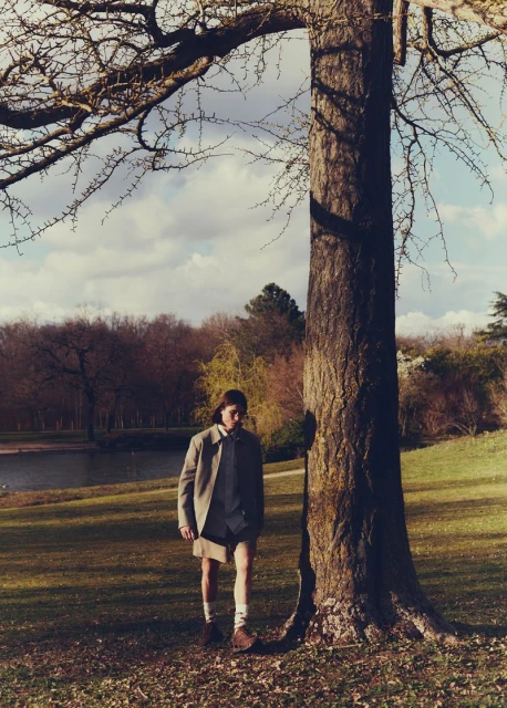 An adult male stands under a large tree in a park-like setting with a pond and trees in the background. He wears a blazer and shorts.