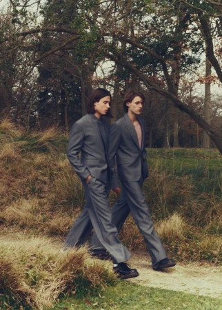 Two male fashion models in grey suits walk on a path through a wooded park setting with bare trees and natural light in an editorial style.