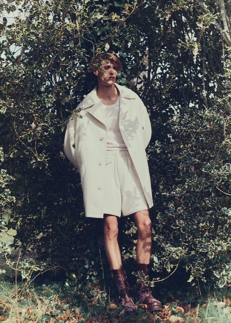 Male fashion model in a cream coat, shorts, and combat boots poses against a lush green leafy backdrop in an outdoor fashion editorial.