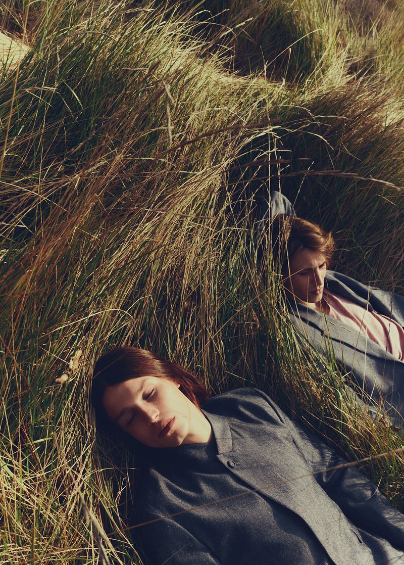 Two adults in suits lie amongst tall, dry grass in a close-up shot at eye-level, bathed in soft, natural lighting, evoking a serene mood.