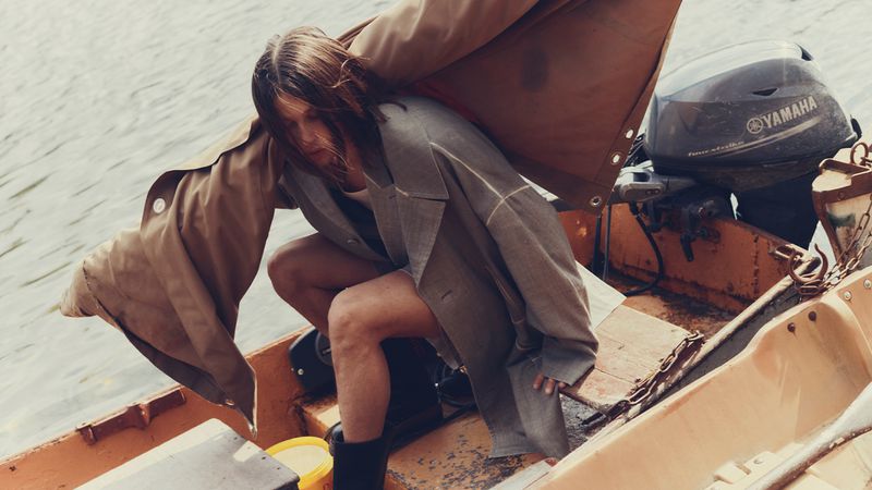 Woman in oversized blazer squats in boat on lake. Taupe and yellow tones with Yamaha motor in background. Fashion editorial style.