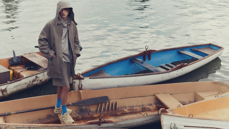 A young adult male stands on a weathered boat wearing a hooded jacket and shorts, with water and other boats as the backdrop.