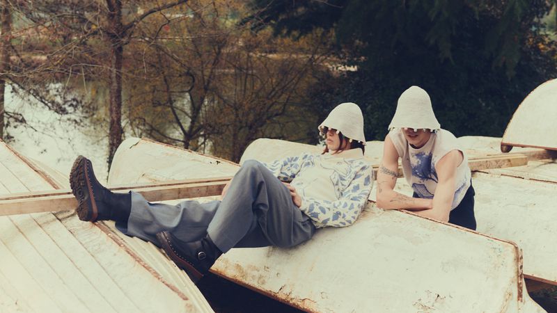 Two people in matching hats pose among rustic boats, set against a backdrop of trees and a calm lake, creating a fashion editorial image.