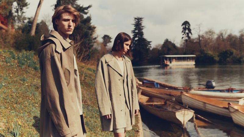 Two people in trench coats stand near boats by a lake on an overcast day, trees in the background, fashion photography.