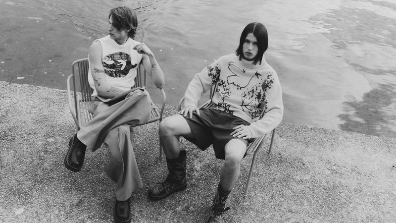 Two male models sit on metal chairs outdoors near rippled water. One wears a tank top and trousers; the other a sweater and shorts.