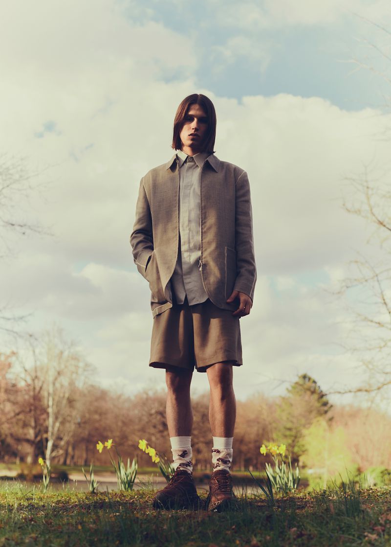 Full-shot of a young adult male standing in a park. He wears a blazer, shorts, and patterned socks near daffodils and trees.