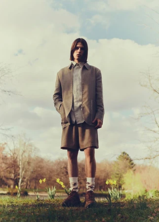 Full-shot of a young adult male standing in a park. He wears a blazer, shorts, and patterned socks near daffodils and trees.