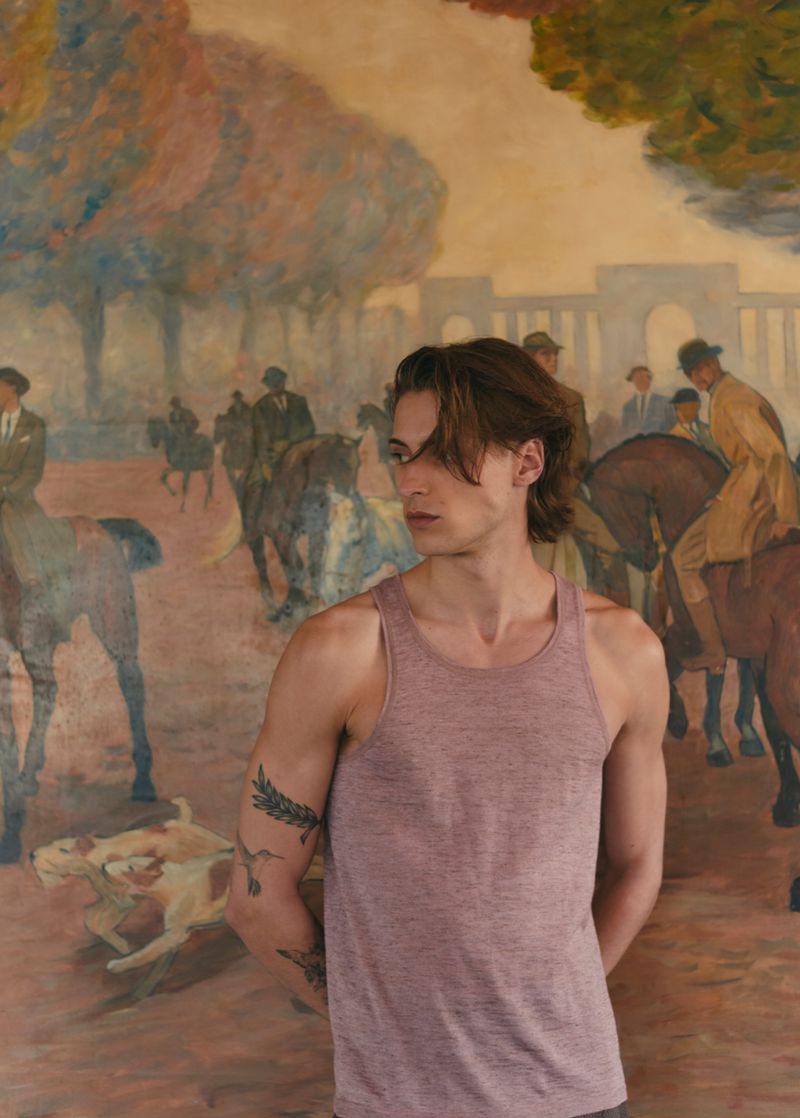 Young adult male in tank top poses against an impressionist painting backdrop featuring horses, dogs, and people in a park.
