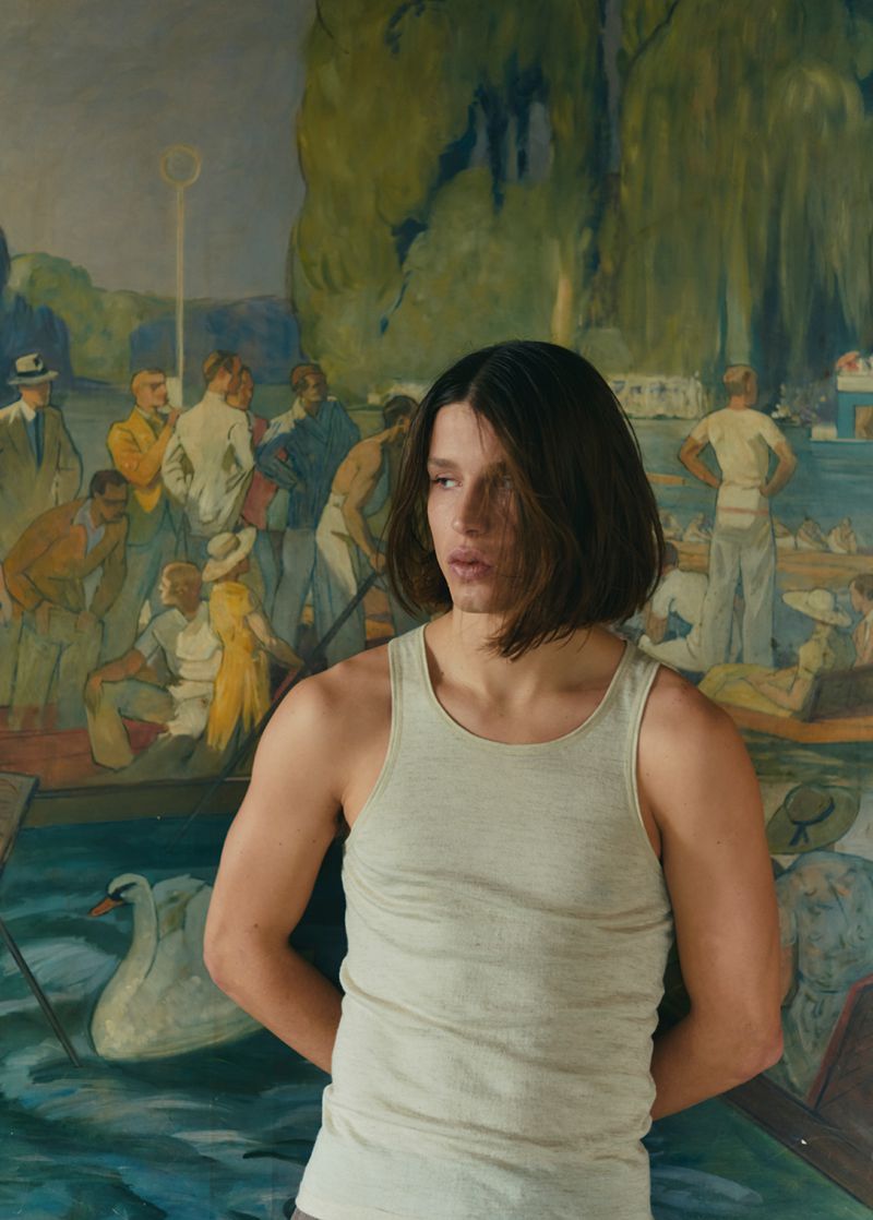 Portrait of a young adult male wearing a tank top, posing against a backdrop of a vintage painting depicting a lakeside scene.