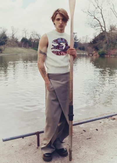 Model holding a wooden oar standing by a lake wearing a white sleeveless graphic knit top and taupe trousers