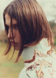 Close-up portrait of a male model with shoulder-length brown hair wearing a light blue turtleneck sweater adorned with fallen autumn leaves.