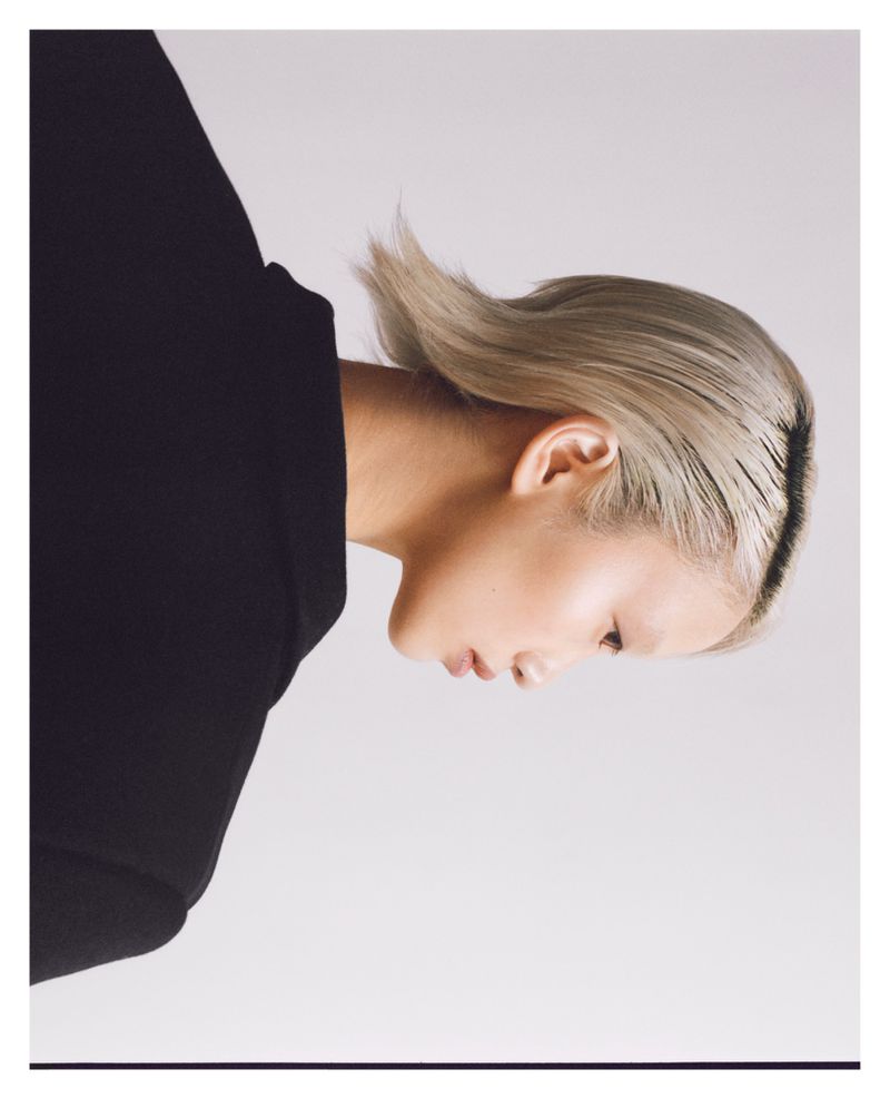 Sideways profile of a person with platinum blonde hair wearing a dark top against a pale grey backdrop in a minimalist composition.