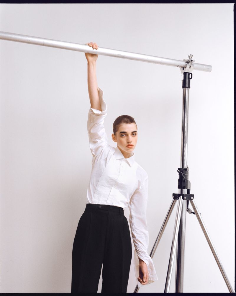 A young woman with shaved head in a white shirt and dark pants is holding onto a horizontal metal bar attached to a photography light stand.