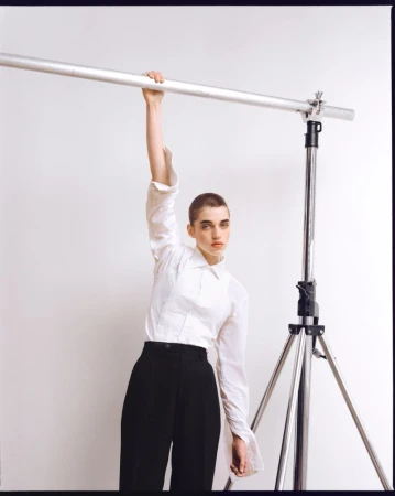 A young woman with shaved head in a white shirt and dark pants is holding onto a horizontal metal bar attached to a photography light stand.