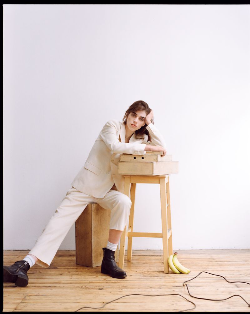 Woman in a beige suit and black boots leans on a wooden stool with stacked boxes in a studio setting, creating a fashionable portrait.