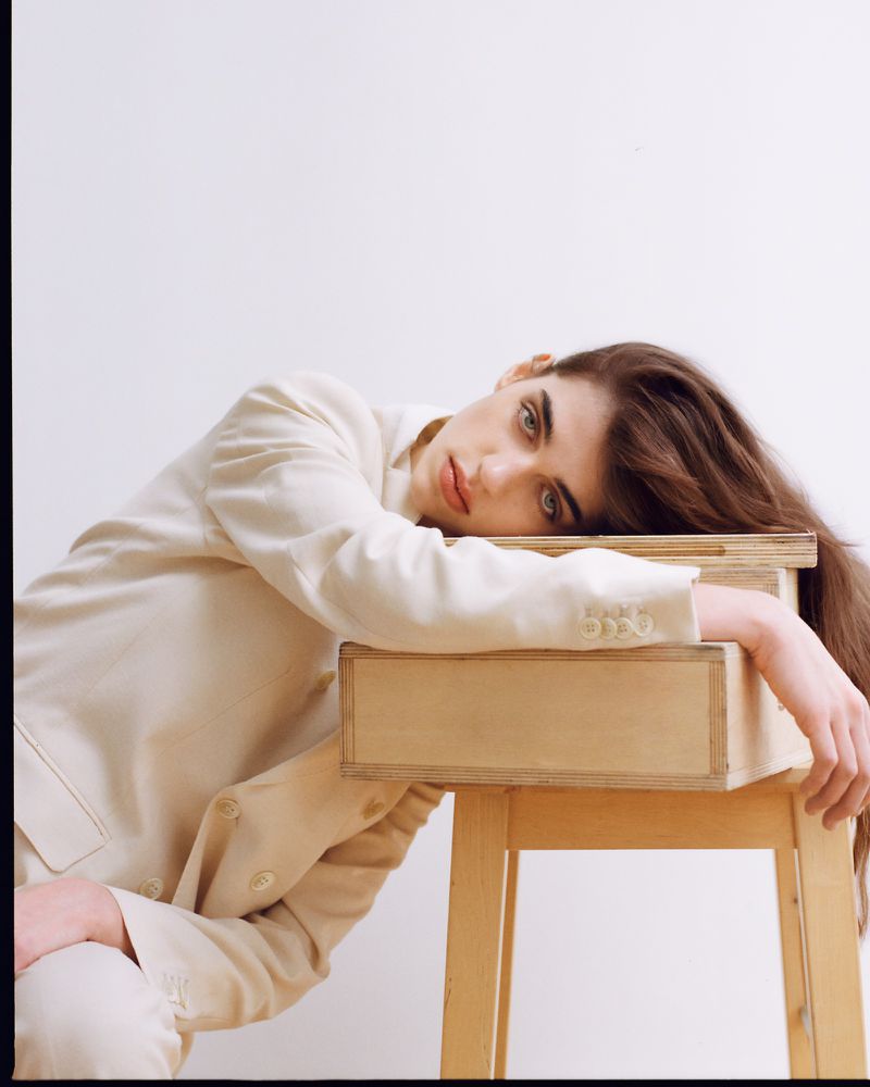 Woman in suit reclines on wooden table in a studio shot. She is fair-skinned with long brown hair and is posed looking at the camera.