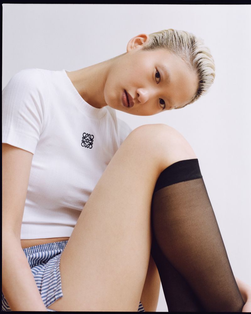 Asian woman with a short blonde haircut poses indoors, wearing a white shirt with a logo, paired with striped shorts and black stockings.