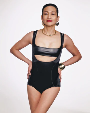 Elegant adult woman in a cutout leotard poses confidently in a studio with gold jewelry, against a bright, neutral backdrop.