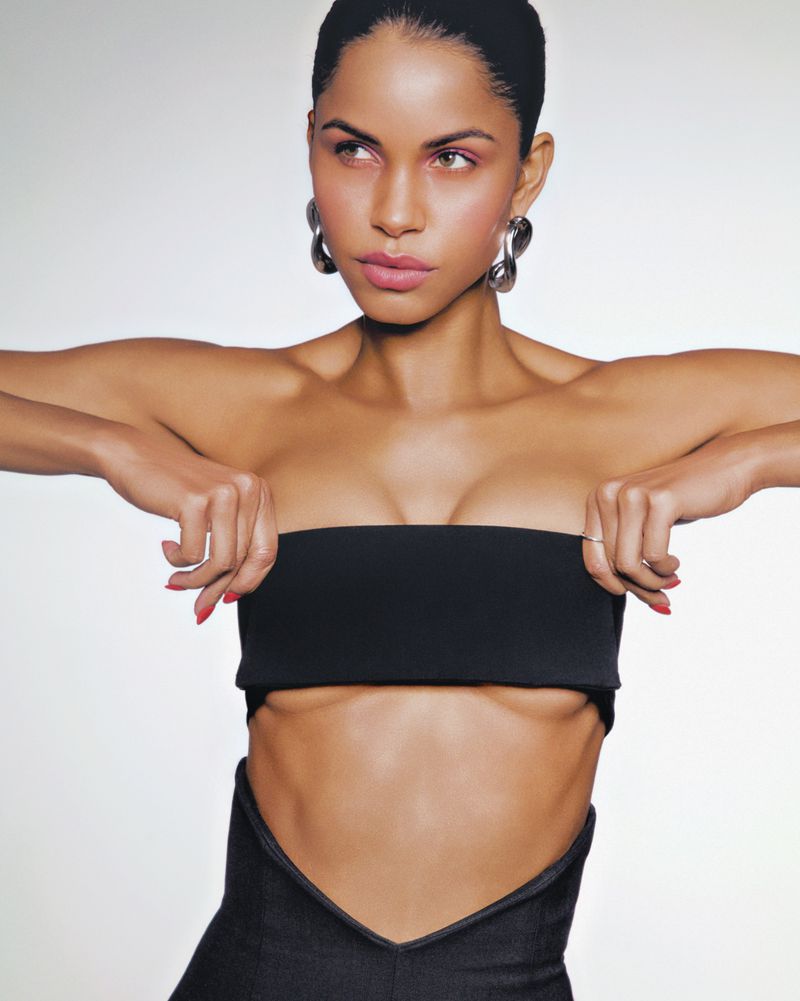 Portrait of a woman in a black tube top and dark jeans against a white background, wearing hoop earrings and red nail polish.