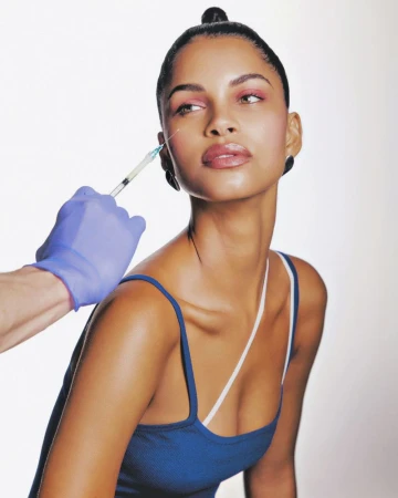 Close-up of a woman receiving a cosmetic injection near her eye by a medical professional wearing blue medical gloves in a studio.