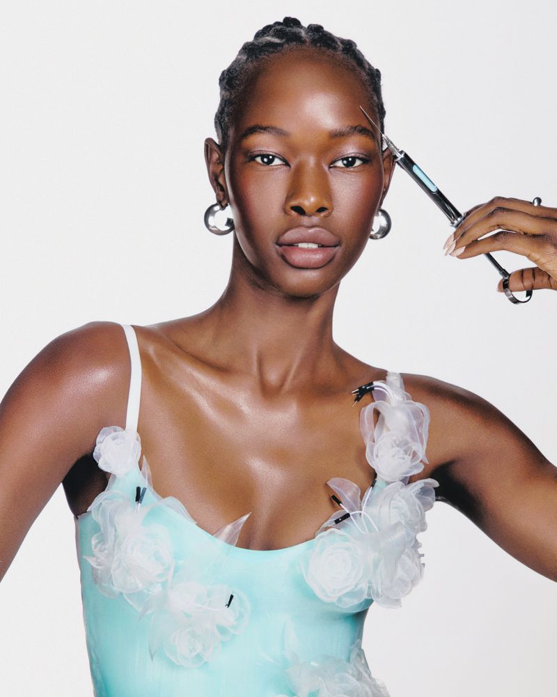 Portrait of a young Black woman in a blue dress adorned with white flowers, getting her eyebrow shaped with small scissors.