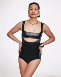 A woman wearing a black cut-out swimsuit and gold statement jewelry poses against a clean white background.