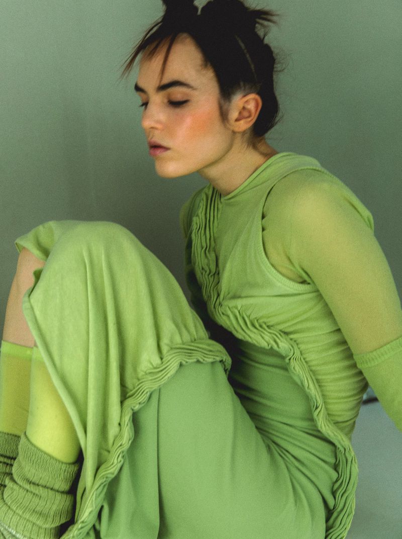 Woman in green dress seated against a green wall. She has brown hair in a top knot and a neutral expression in a close-up studio shot.