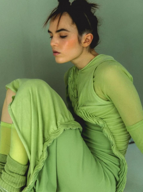 Woman in green dress seated against a green wall. She has brown hair in a top knot and a neutral expression in a close-up studio shot.