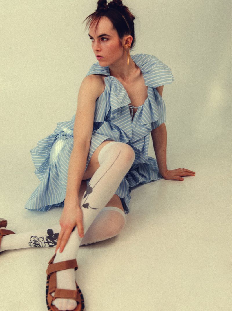 A young adult female poses indoors in a blue and white dress, knee-high socks with graphics, and sandals against a plain backdrop.