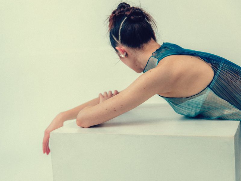 Back view of a slender adult female resting on a geometric white block, wearing a sheer blue dress against a neutral background.
