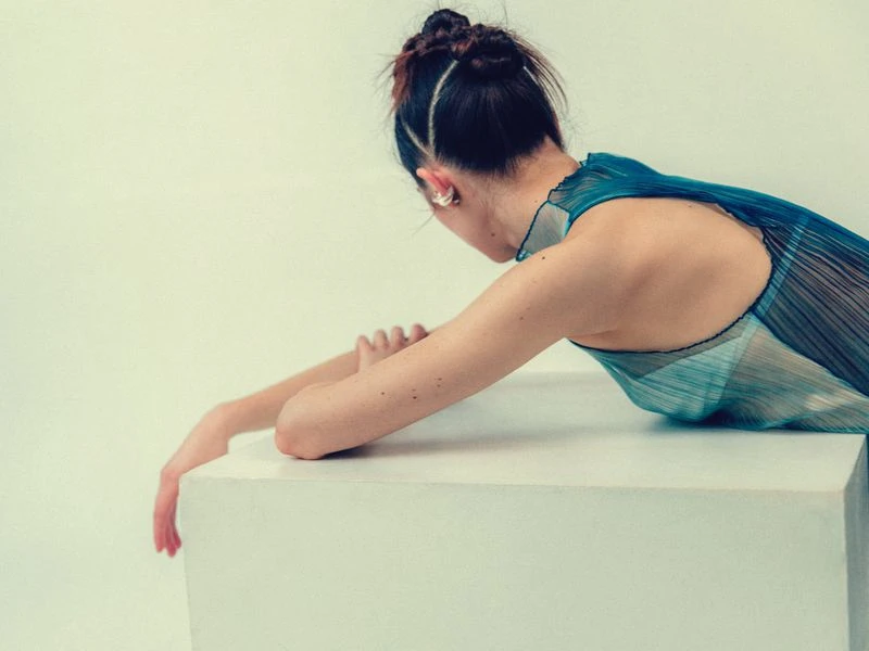 Back view of a slender adult female resting on a geometric white block, wearing a sheer blue dress against a neutral background.