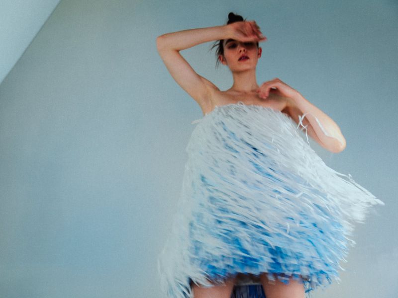 Model wearing a textured white and blue fringe dress posing with an arm raised against a plain wall