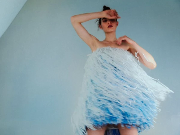 Model wearing a textured white and blue fringe dress posing with an arm raised against a plain wall