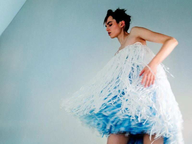 Woman in blue-and-white dress with fringe posing against light blue background in studio setting, fashion portrait.