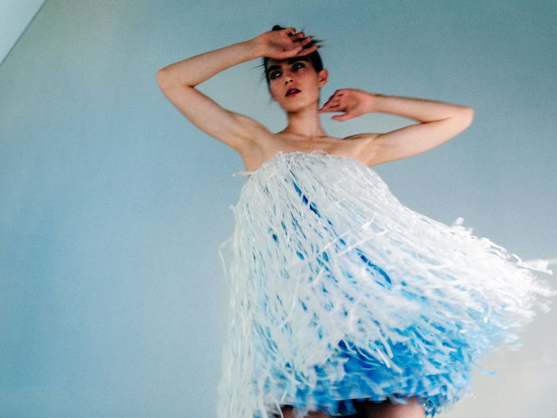 Woman in strapless blue and white fringe dress poses in front of a blue background, hands above her head, looking at camera.