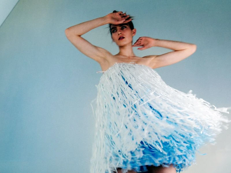 Woman in strapless blue and white fringe dress poses in front of a blue background, hands above her head, looking at camera.