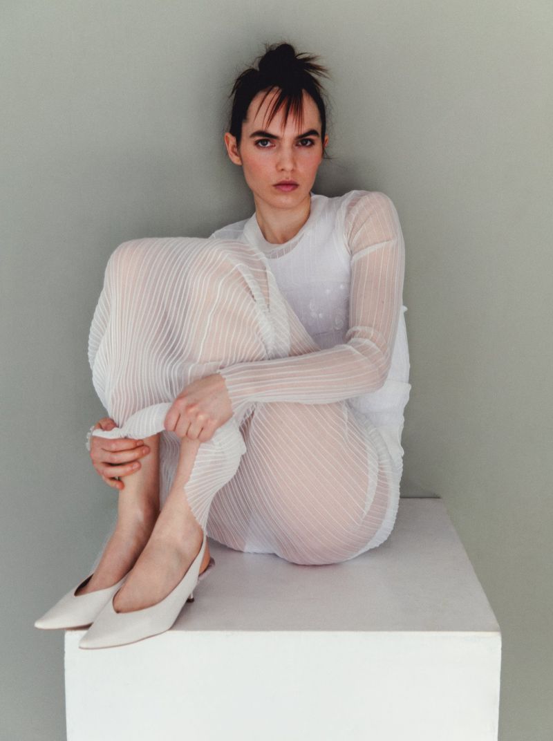 A fair-skinned adult woman sits on a white cube, wearing a white mesh dress and white shoes, in a simple indoor setting against a grey wall.