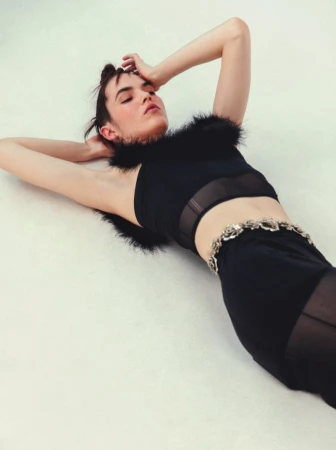 Model lying on a white background wearing a black feather-trimmed top and dark skirt with an ornate belt.