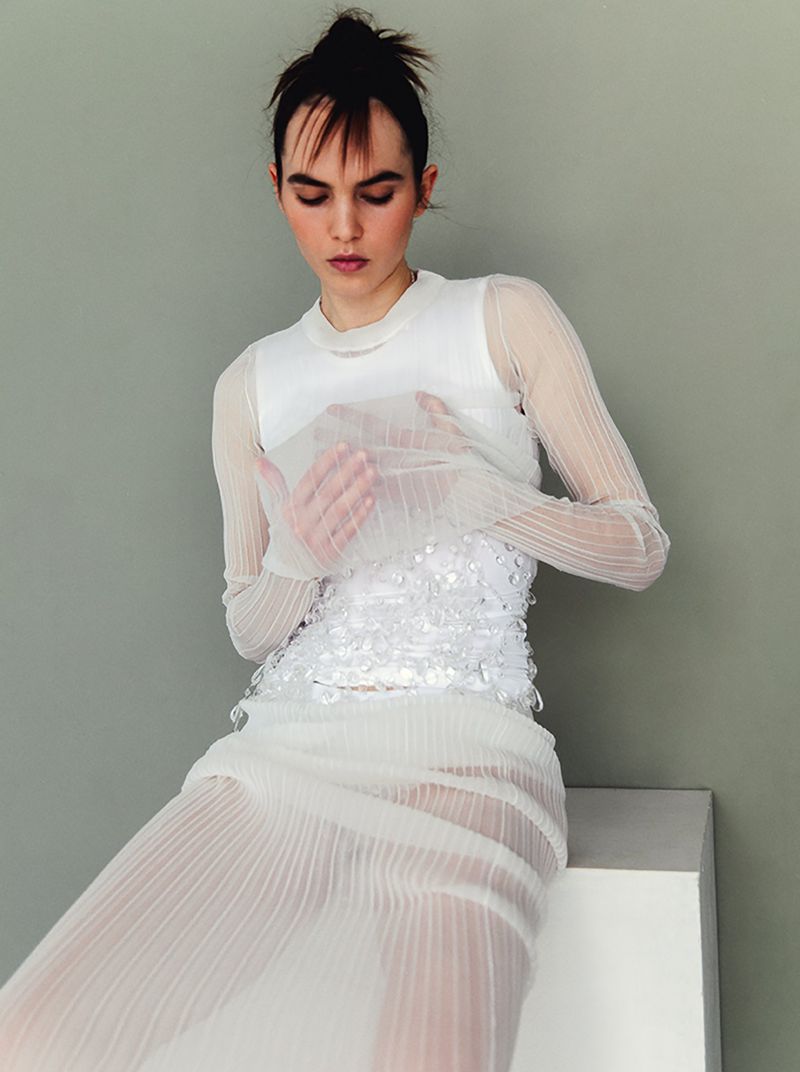 Woman in sheer white dress sits on a white block looking down. Soft studio light and neutral grey background.