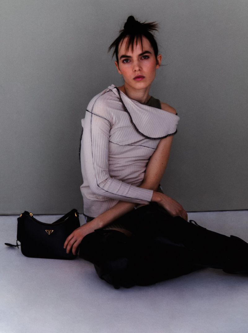 A stylish woman poses in a beige top and black leggings, seated next to a black Prada handbag in a fashion editorial shot.