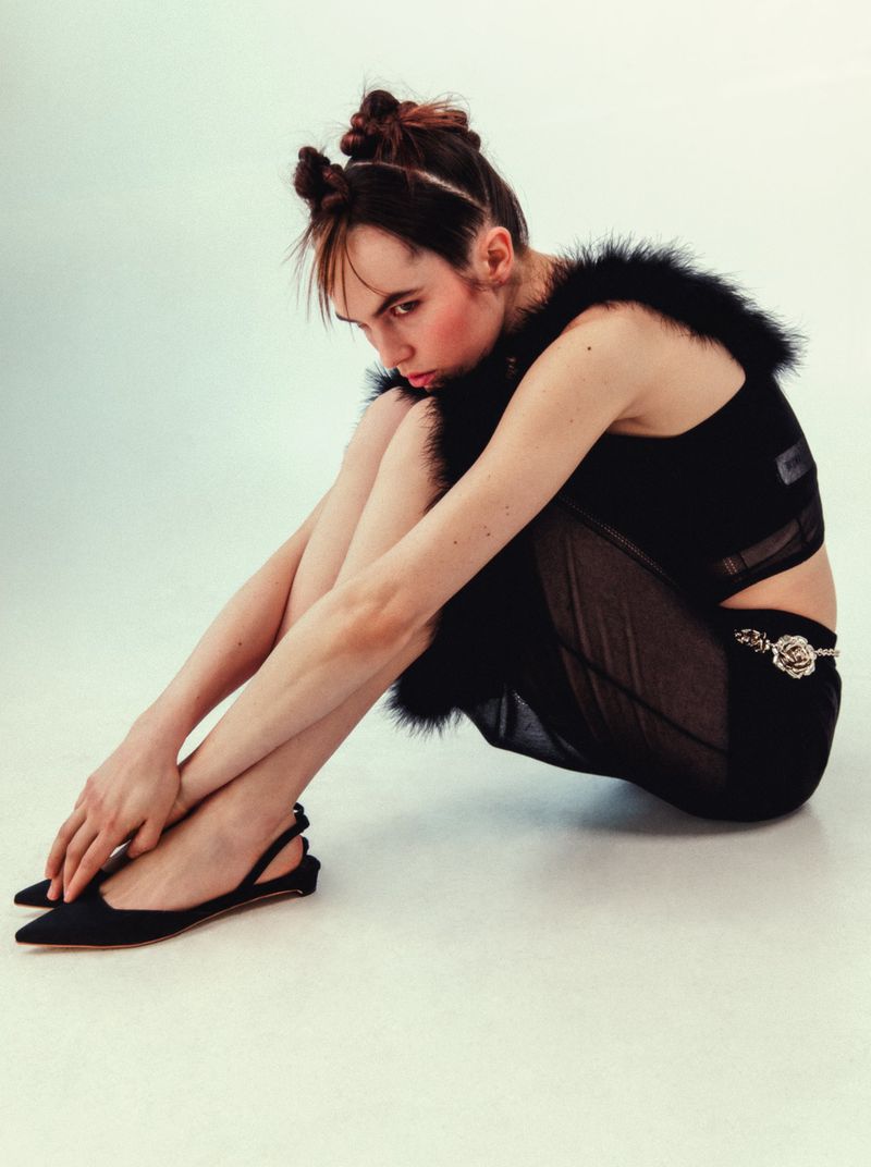 Seated young woman in black outfit with space buns poses in a studio. Dark top with fur trim and sheer skirt paired with slingback heels.