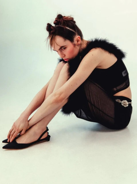 Seated young woman in black outfit with space buns poses in a studio. Dark top with fur trim and sheer skirt paired with slingback heels.